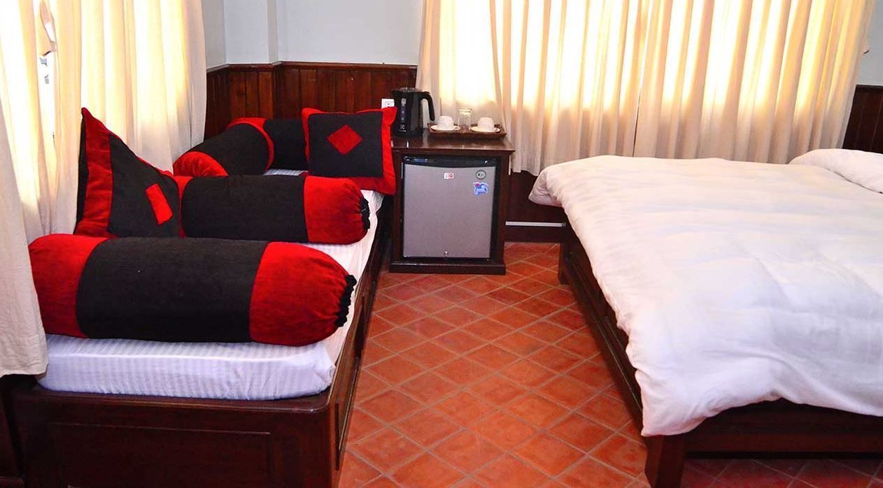A hotel room with two beds, one with red and black bedding and the other with white bedding. There is a small table with a coffee maker and cups, and a refrigerator.