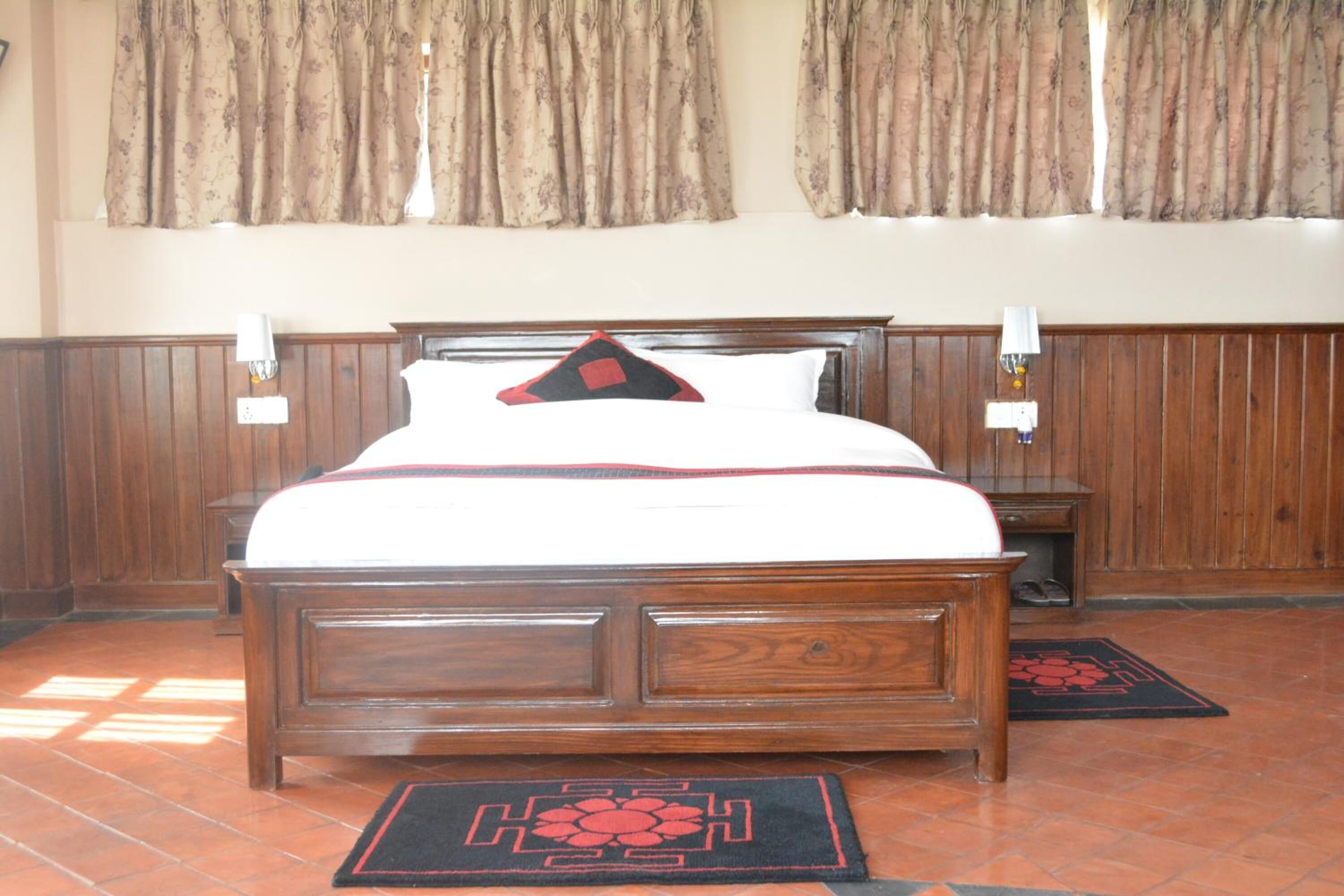 A wooden four-poster bed with a white bedspread and a red and black pillow. There are two bedside tables with lamps, a wooden floor, and a window with curtains.
