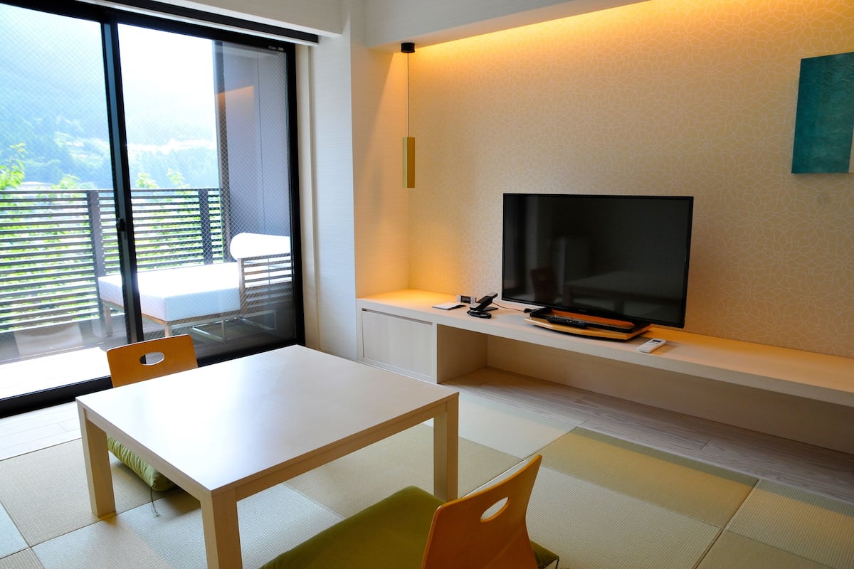 Una habitación moderna con un ventanal grande, un televisor de pantalla plana sobre un soporte blanco, una mesa de madera y un cojín verde.
