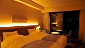Habitación de hotel con dos camas, una mesita de noche, una silla, una pequeña mesa y vista a la ciudad de noche.
