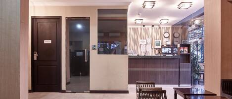 Un hall de hotel con recepción, puerta de cristal de acceso y zona de estar con muebles de madera.