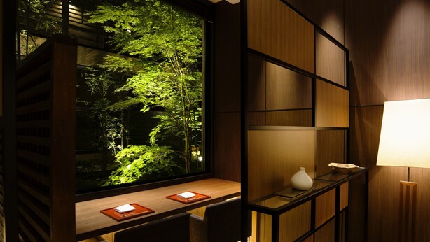 Un intérieur moderne avec une grande fenêtre donnant sur une forêt, un bureau en bois avec deux chaises et une étagère au design minimaliste.