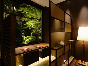 Un interior moderno con un ventanal grande que da a un bosque, un escritorio de madera con dos sillas y una estantería minimalista.
