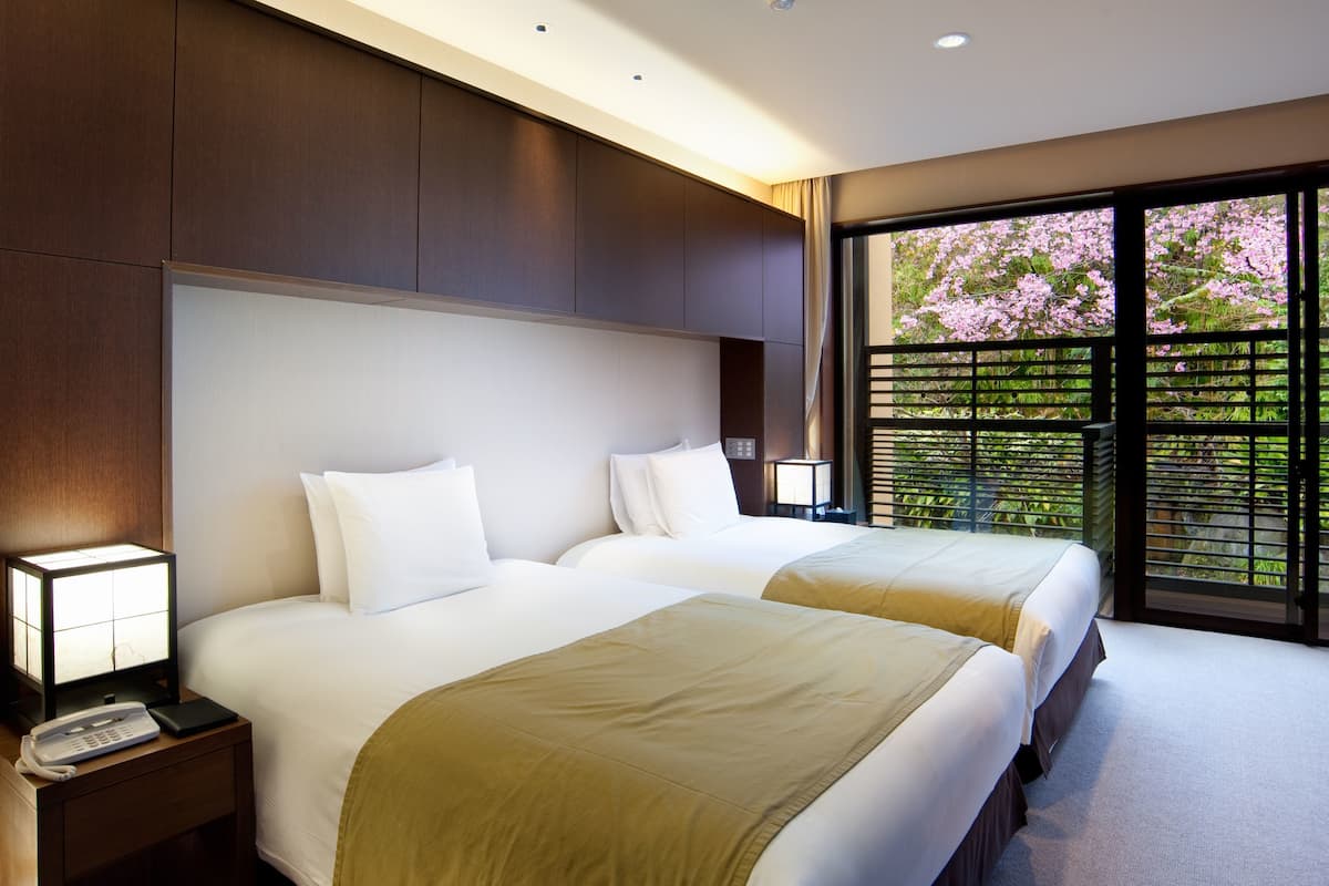 A hotel room with two beds, a nightstand with a phone, a lamp, and a view of cherry blossoms through a window.