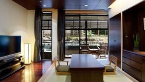 Une chambre d’hôtel moderne dotée d’une grande télévision à écran plat, d’une table à manger en bois avec des chaises, d’un canapé et offrant une vue sur un jardin grâce à de grandes fenêtres.