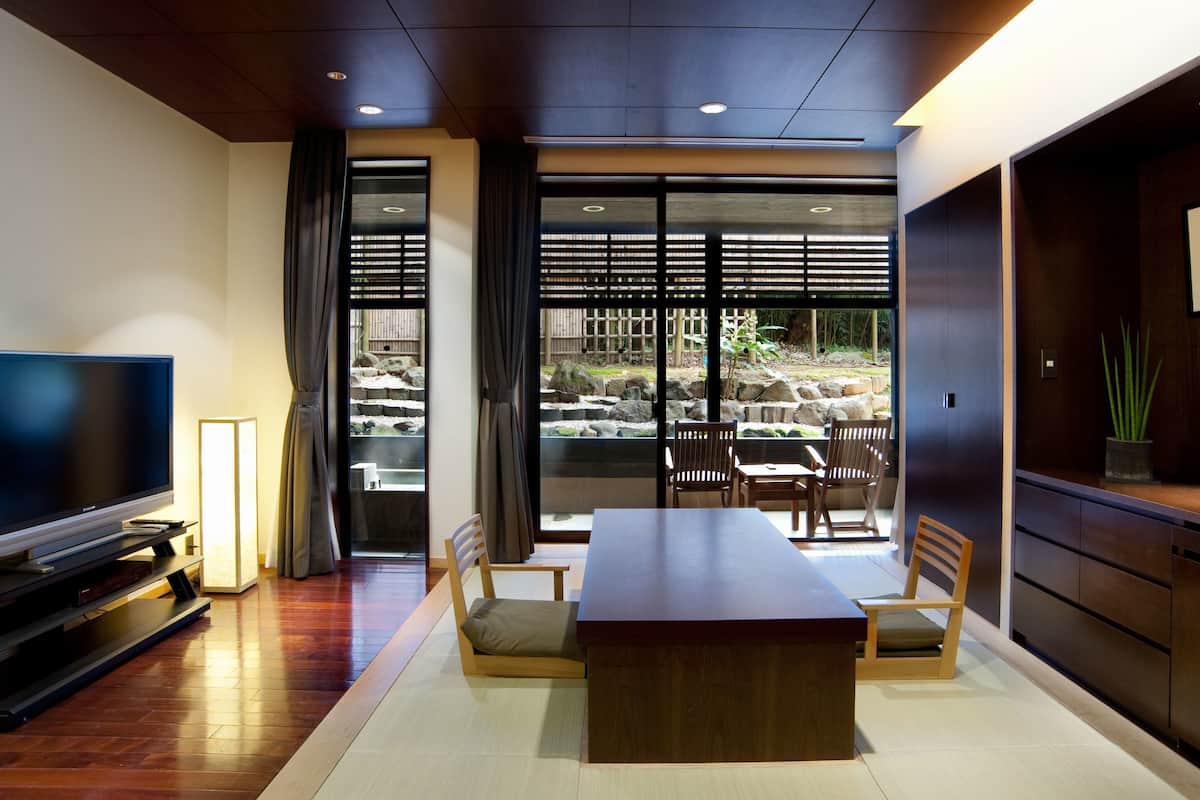 A modern hotel room with a large flat-screen TV, a wooden dining table with chairs, a sofa, and a view of a garden through large windows.