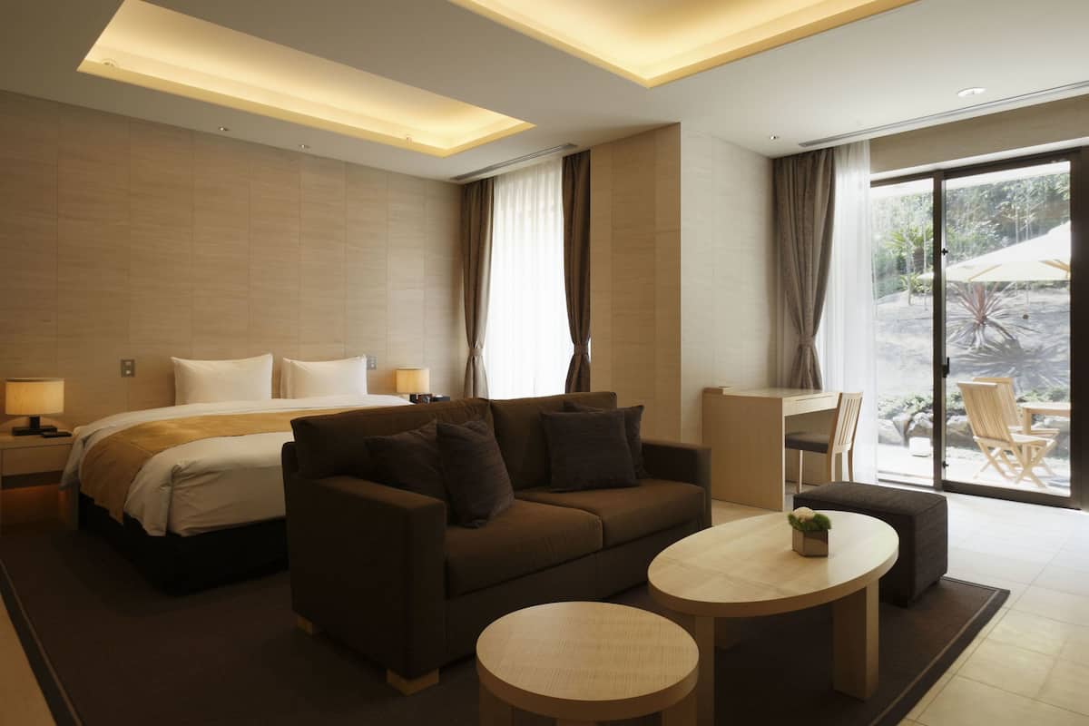 A modern hotel room with a bed, a sofa, a desk, and a view of the outdoors.