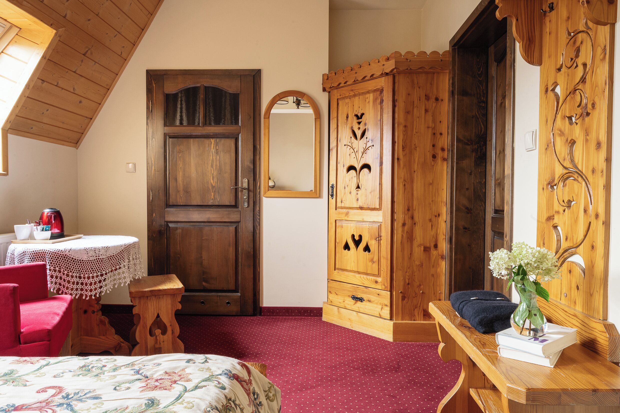 Um quarto aconchegante com um guarda-roupa de madeira, um espelho, uma mesinha com uma cadeira vermelha e uma cama com colcha florida.
