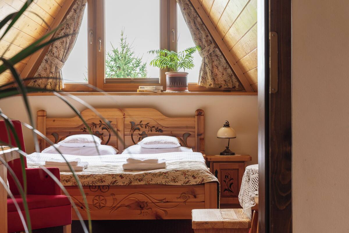 A bedroom with a wooden bed, a bedside table with a lamp, a window with a view of trees, and a red chair.