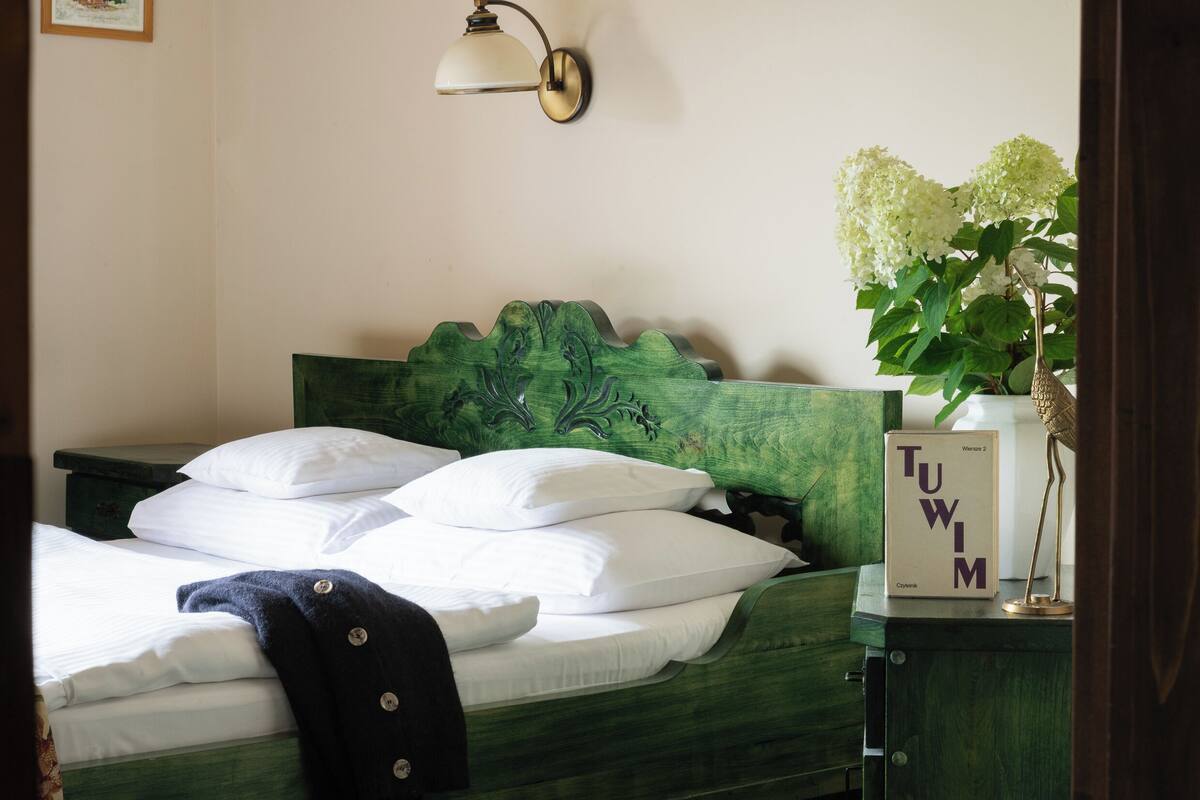 A neatly made bed with white pillows and a dark throw blanket, a green headboard with a carved design, a wall-mounted lamp, and a vase with white flowers on a nightstand.