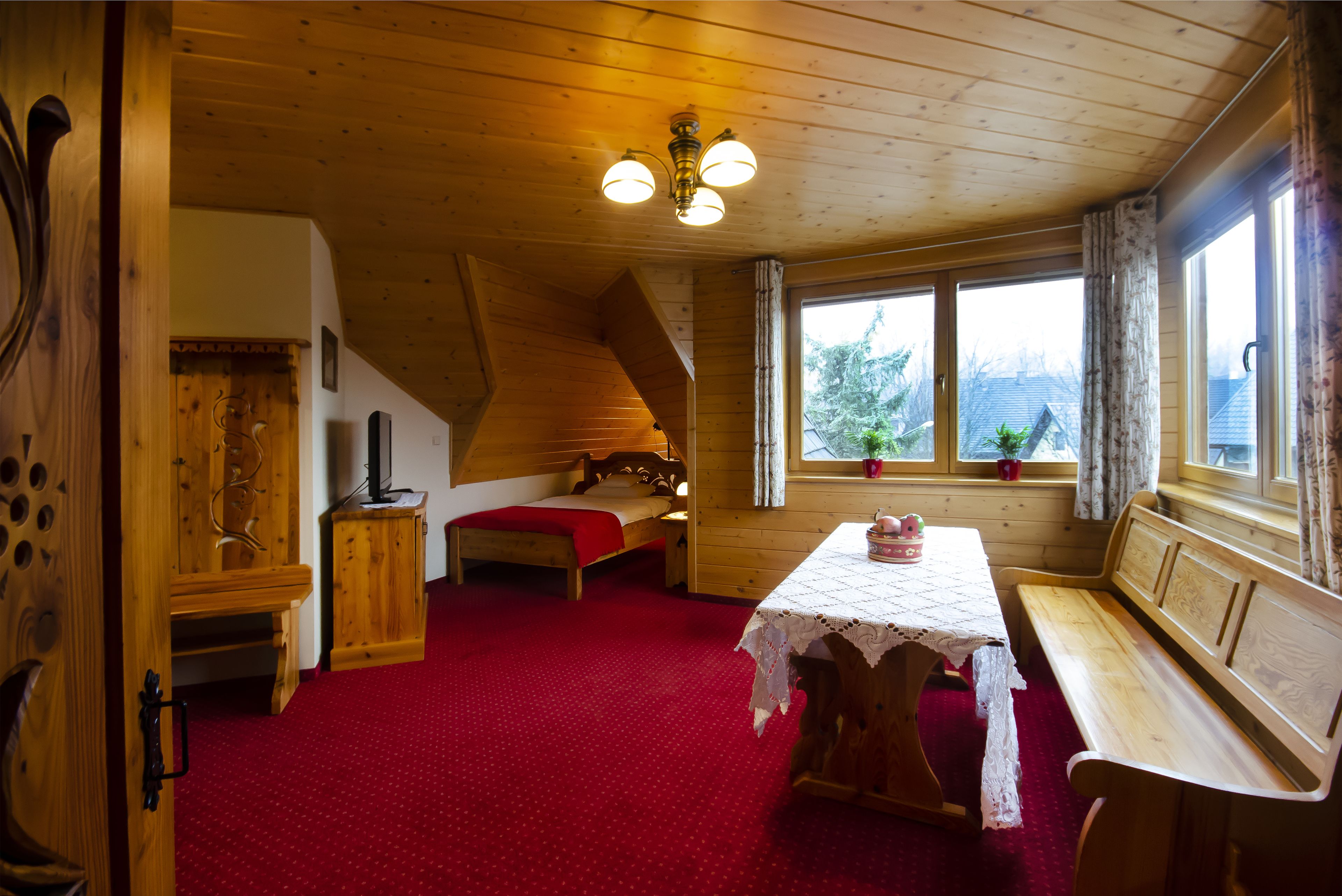 A cozy room with a wooden ceiling, a bed, a table with a lace tablecloth, and a bench.