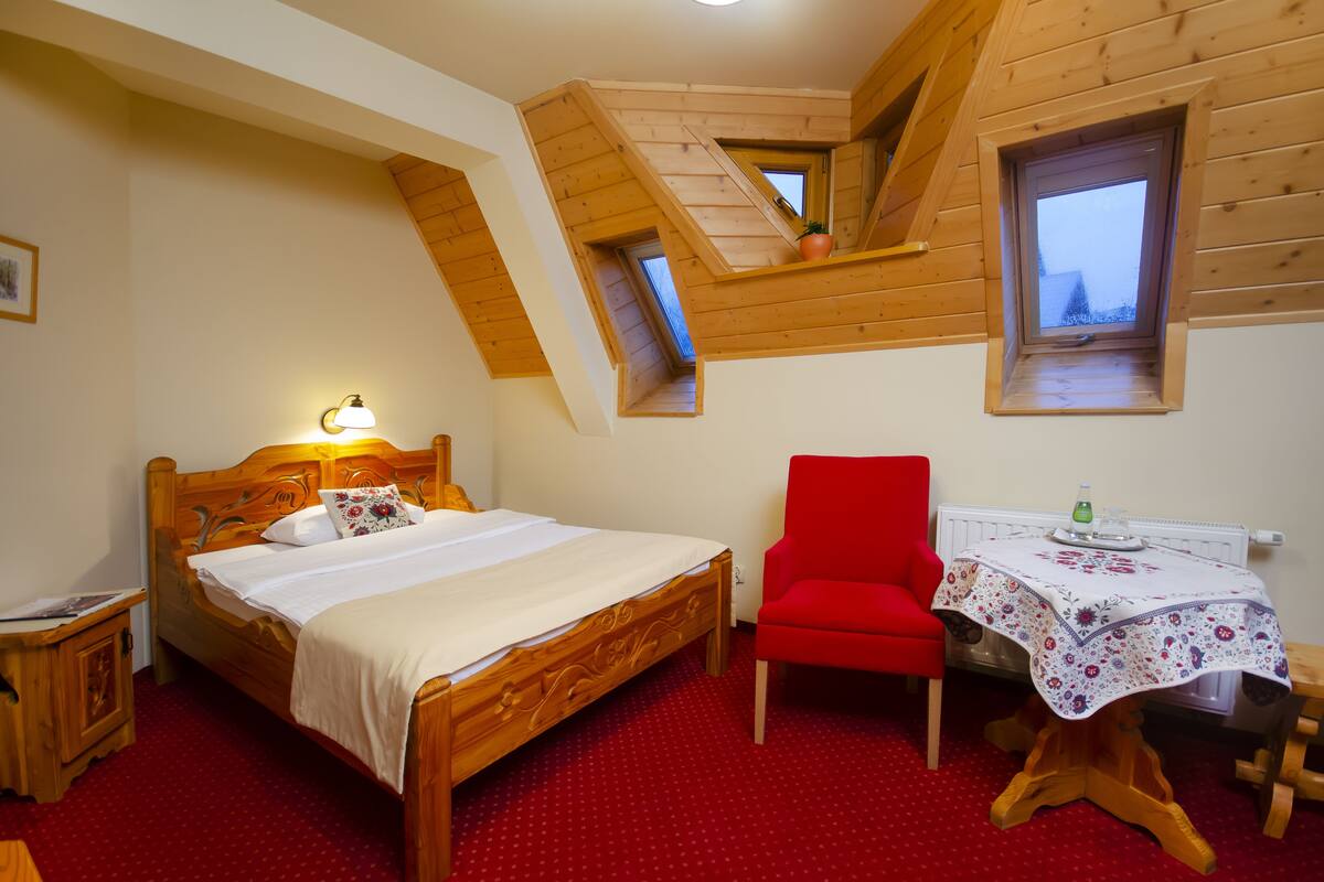 A bedroom with a wooden bed, a red chair, a small table with a tablecloth, and a window with a view.