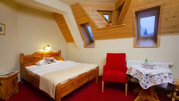 A bedroom with a wooden bed, a red chair, a small table with a tablecloth, and a window with a view.