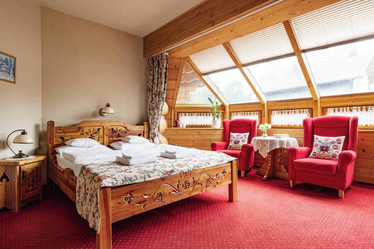 A wooden-framed room with a bed, two red armchairs, a small table, and a window with a view.