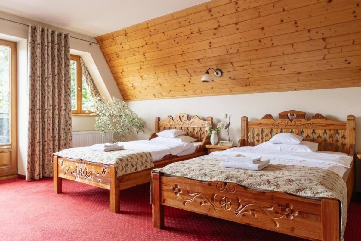 A room with two wooden beds, a red carpet, a wooden ceiling, and a window with curtains.