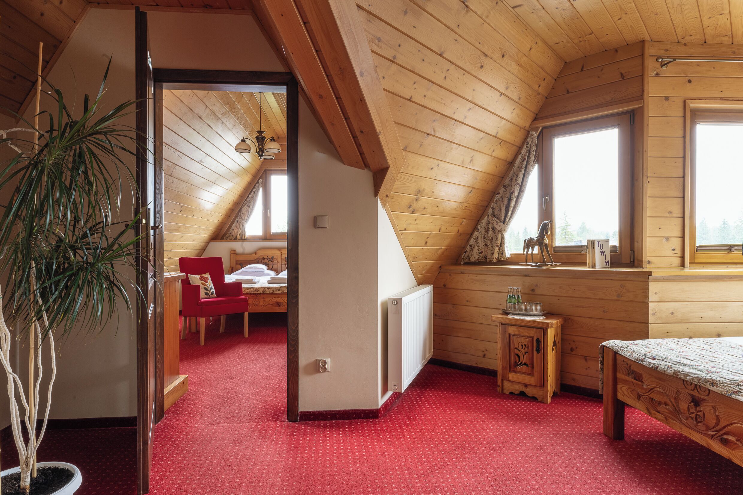 A cozy attic bedroom with wooden beams, a red carpet, a bed, a small table, and a window with a view.