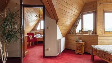 A cozy attic bedroom with wooden beams, a red carpet, a bed, a small table, and a window with a view.