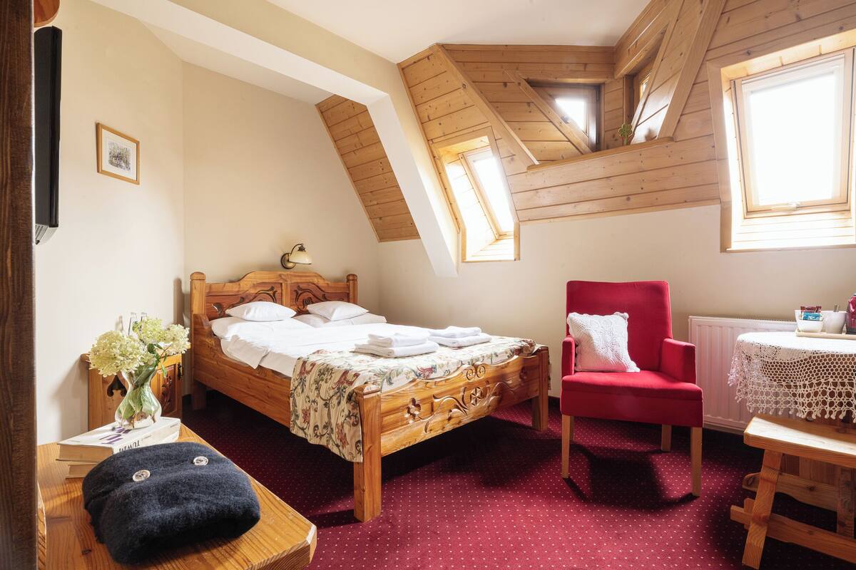 A bedroom with a wooden bed, a red armchair, a small table with a lace tablecloth, and a window with wooden beams.