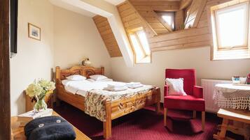 A bedroom with a wooden bed, a red armchair, a small table with a lace tablecloth, and a window with wooden beams.