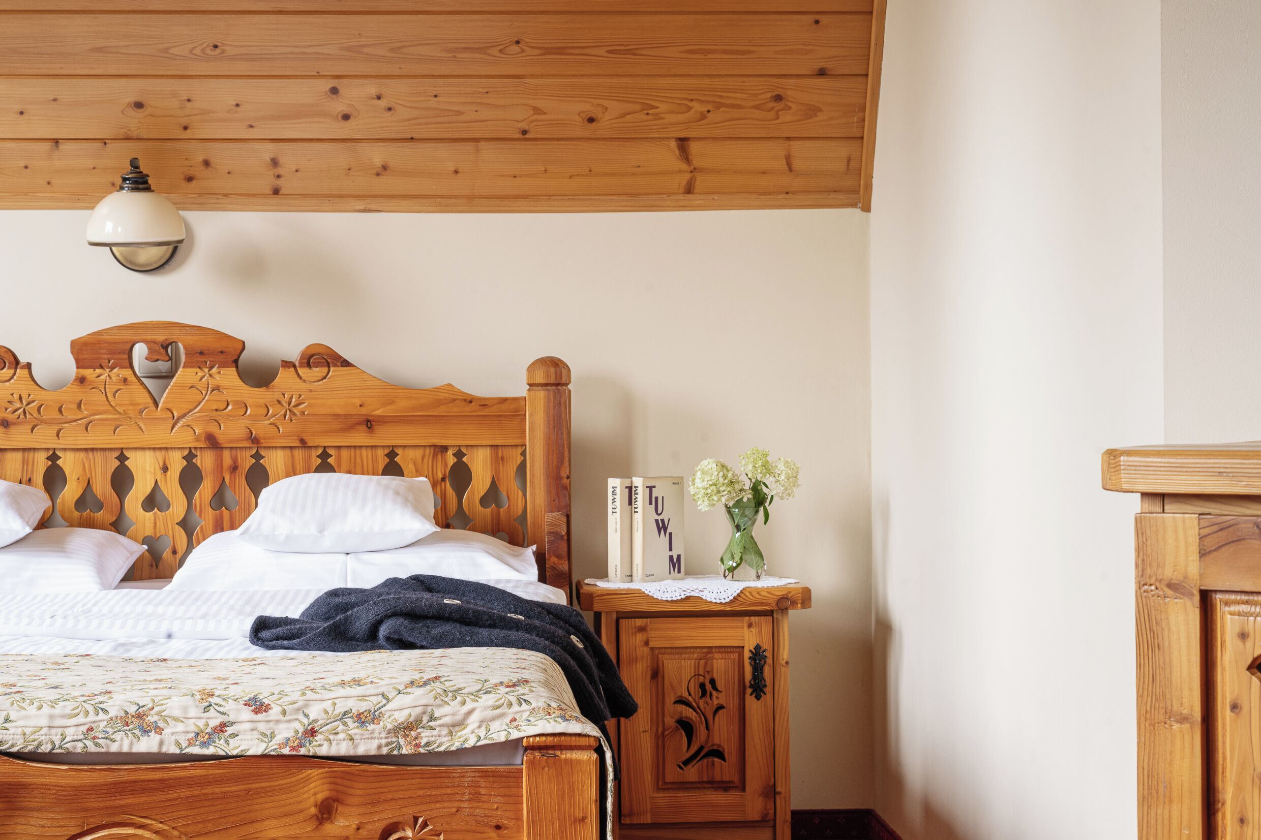 Una cama de madera con cabecera, una mesita de noche con un florero y libros, y una lámpara fijada a la pared.
