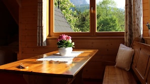 Interior de una cabaña de madera con una mesa de madera, un jarrón blanco con flores y vistas a montañas a través de una ventana.