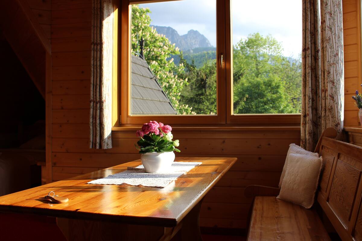 A wooden cabin interior with a wooden table, a white vase with flowers, and a view of mountains through a window.