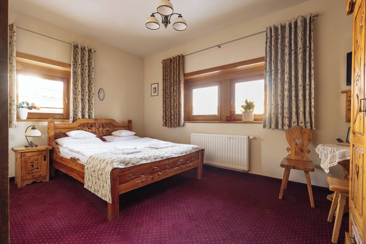 A bedroom with a wooden bed, a nightstand, a chair, a table, and a window with curtains.