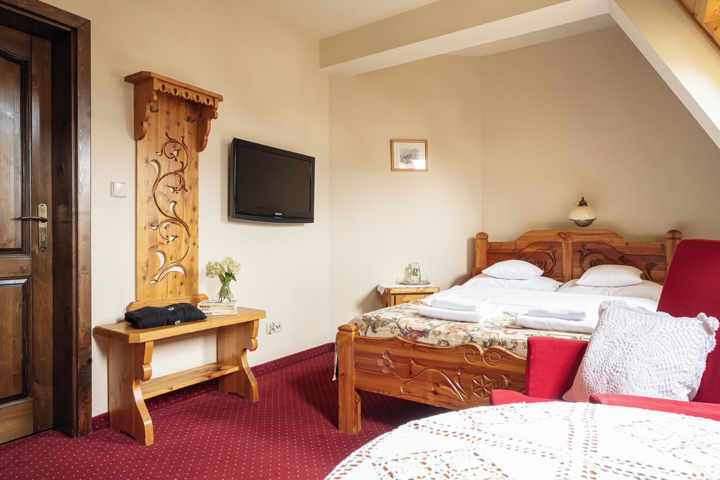 A bedroom with a wooden bed, a red chair, a wooden desk, a television, and a small bedside table.