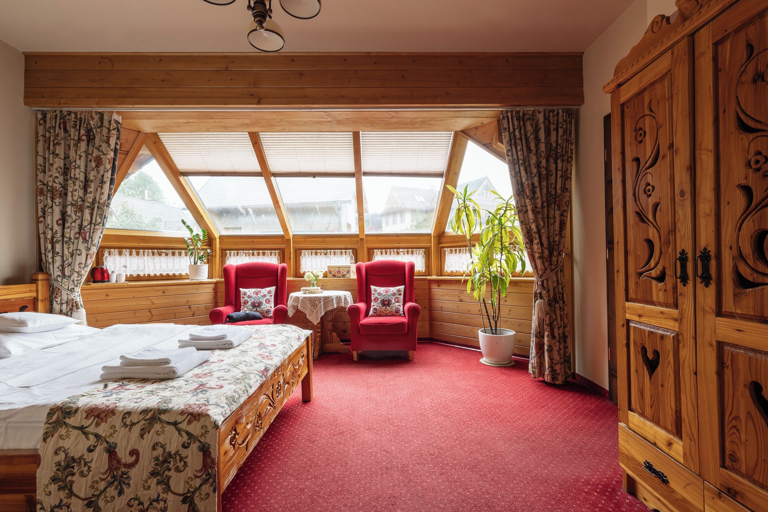 A cozy room with a bed, two red armchairs, a small table with a plant, and a large wooden wardrobe.