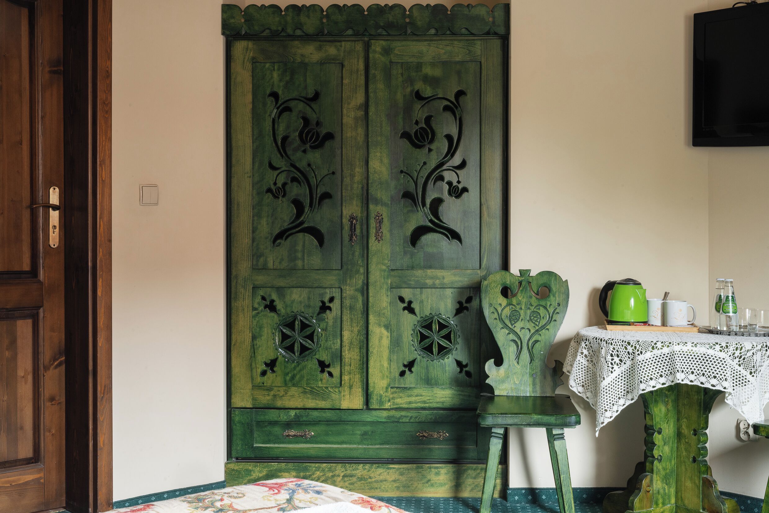 A room with a green wardrobe, a wooden chair, a table with a lace tablecloth, and a television on the wall.