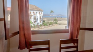 Quarto com uma grande janela com vista para uma praia, uma palmeira e um edifício com telhado de azulejos.
