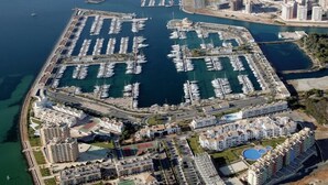Vista aérea de um porto com inúmeros barcos atracados, rodeado por edifícios residenciais e uma estrada costeira.