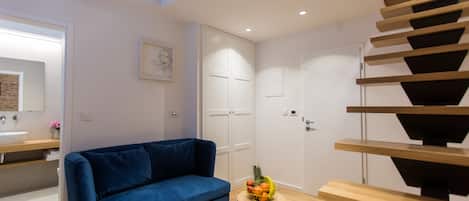 Uma sala de estar moderna com um sofá azul, uma mesa redonda pequena com frutas, um banco de madeira e uma escada.
