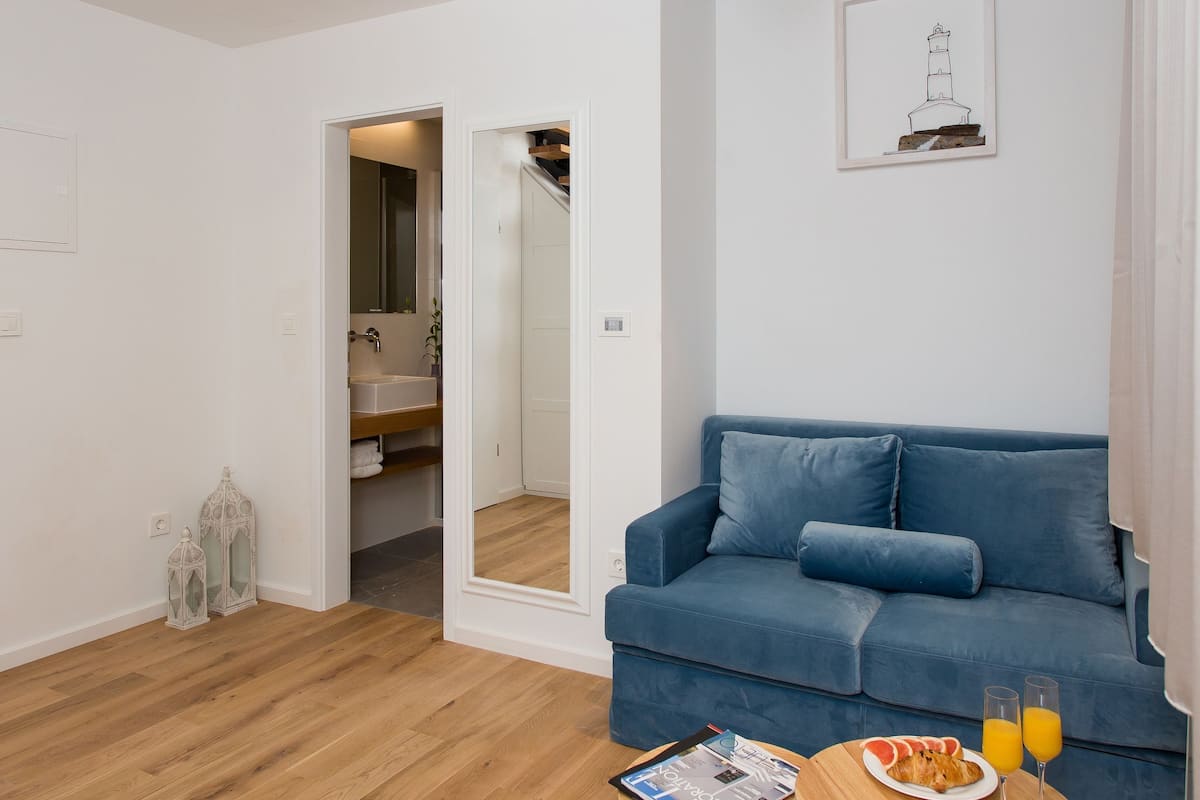 A modern living room with a blue sofa, wooden flooring, a glass staircase, and a dining area with a table and chairs.