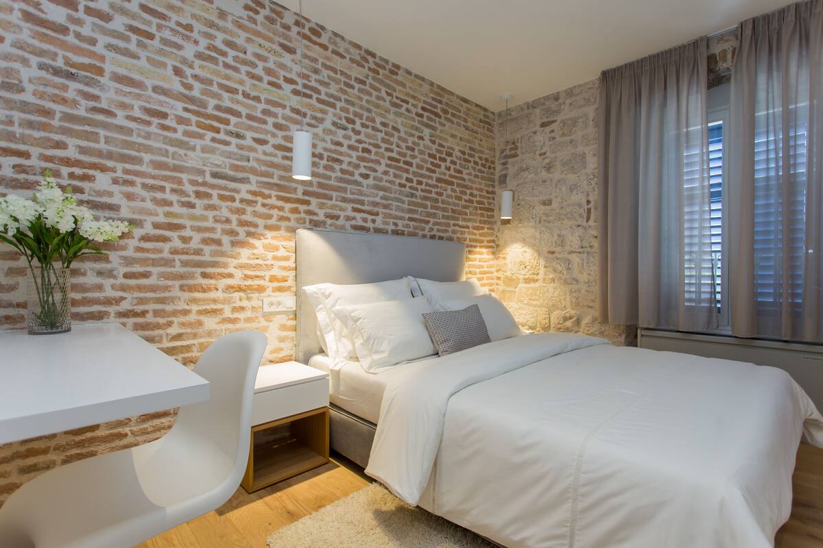 A modern bedroom with a brick wall, a white bed, a bedside table, and a chair.