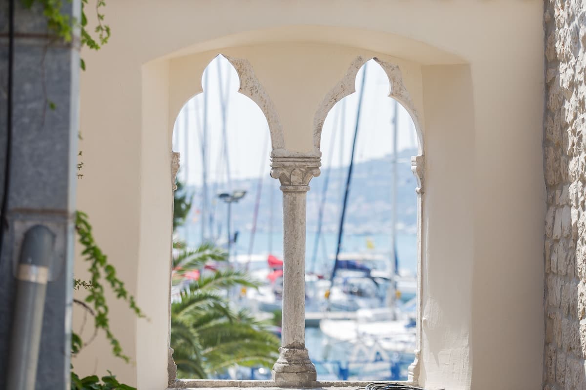 A view through an arched window with a stone balustrade, overlooking a marina with boats and a clear sky.