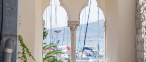 A view through an arched window with a stone balustrade, overlooking a marina with boats and a clear sky.