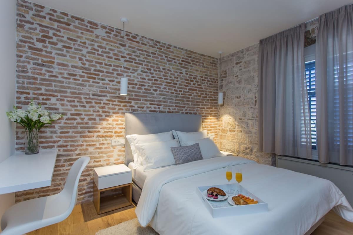 A bedroom with a brick wall, a bed with white bedding, a bedside table, a chair, and a breakfast tray with croissants and juice.