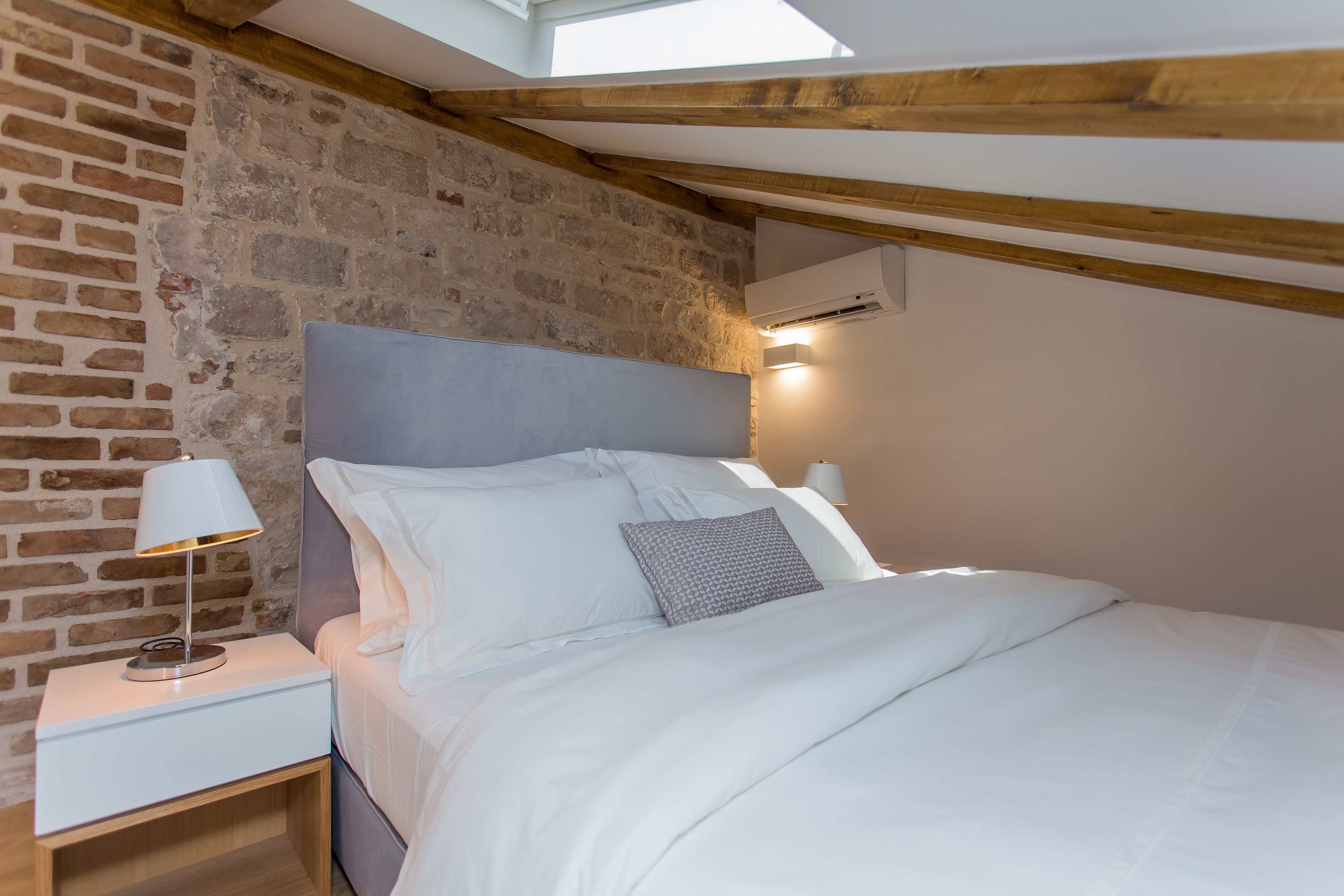 Quarto com parede de tijolos, teto de madeira, cama com roupa de cama branca e mesa de cabeceira com abajur.