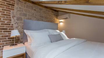 Quarto com parede de tijolos, teto de madeira, cama com roupa de cama branca e mesa de cabeceira com abajur.