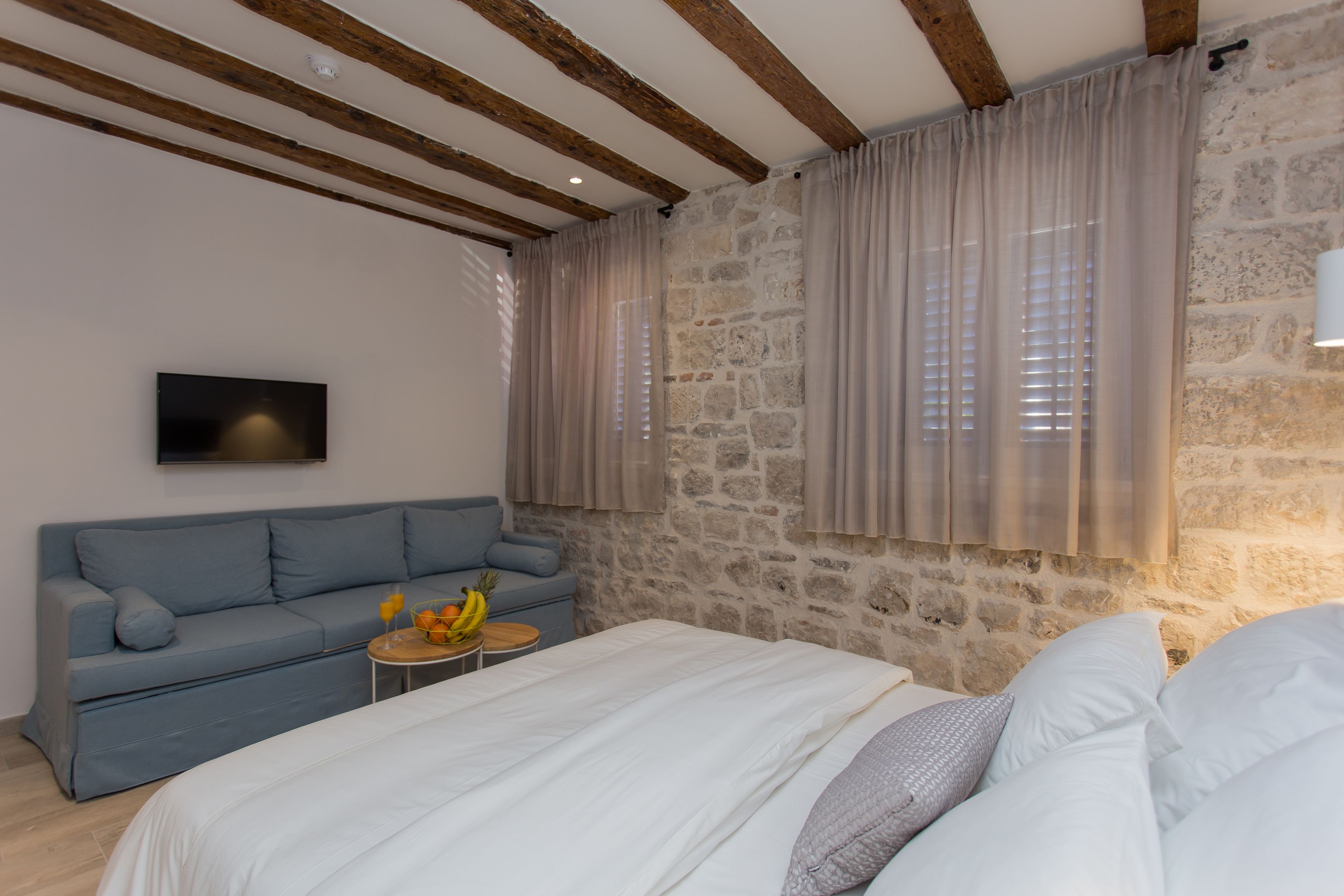 Een slaapkamer met een stenen muur, een houten plafond, een bed met wit beddengoed, een blauwe bank en een kleine tafel met fruit.