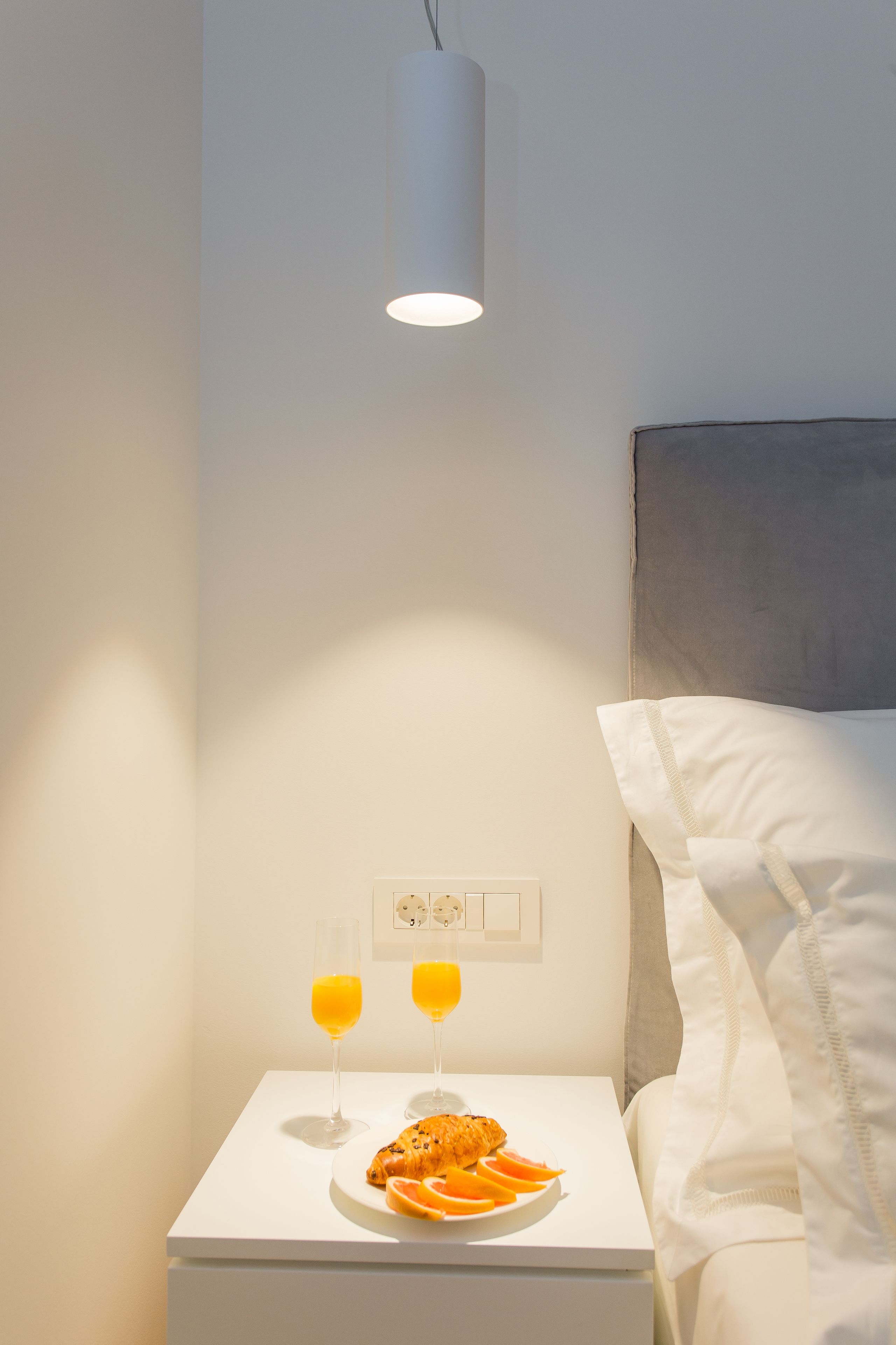A neatly made bed with white linens and a headboard, a bedside table with a plate of food and two glasses of orange juice, and a wall socket with a power outlet.