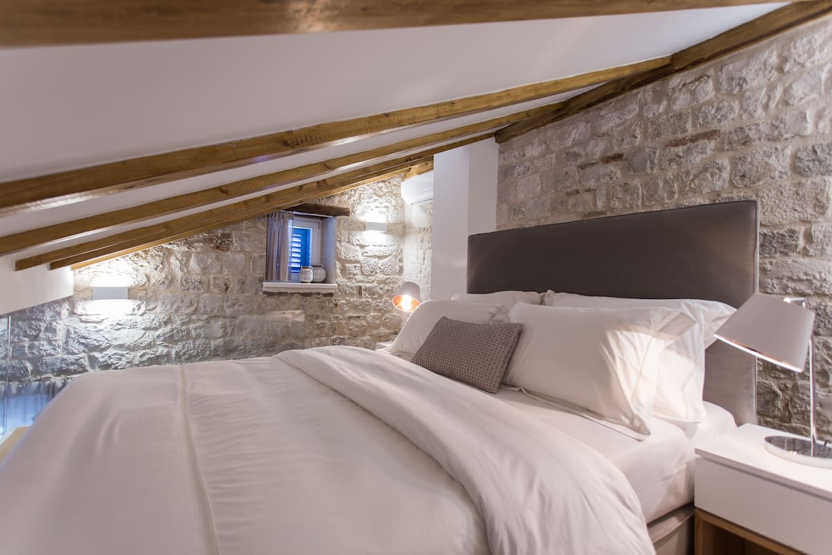 A bedroom with a stone wall, a wooden ceiling, a bed with white bedding, and a nightstand with a lamp.