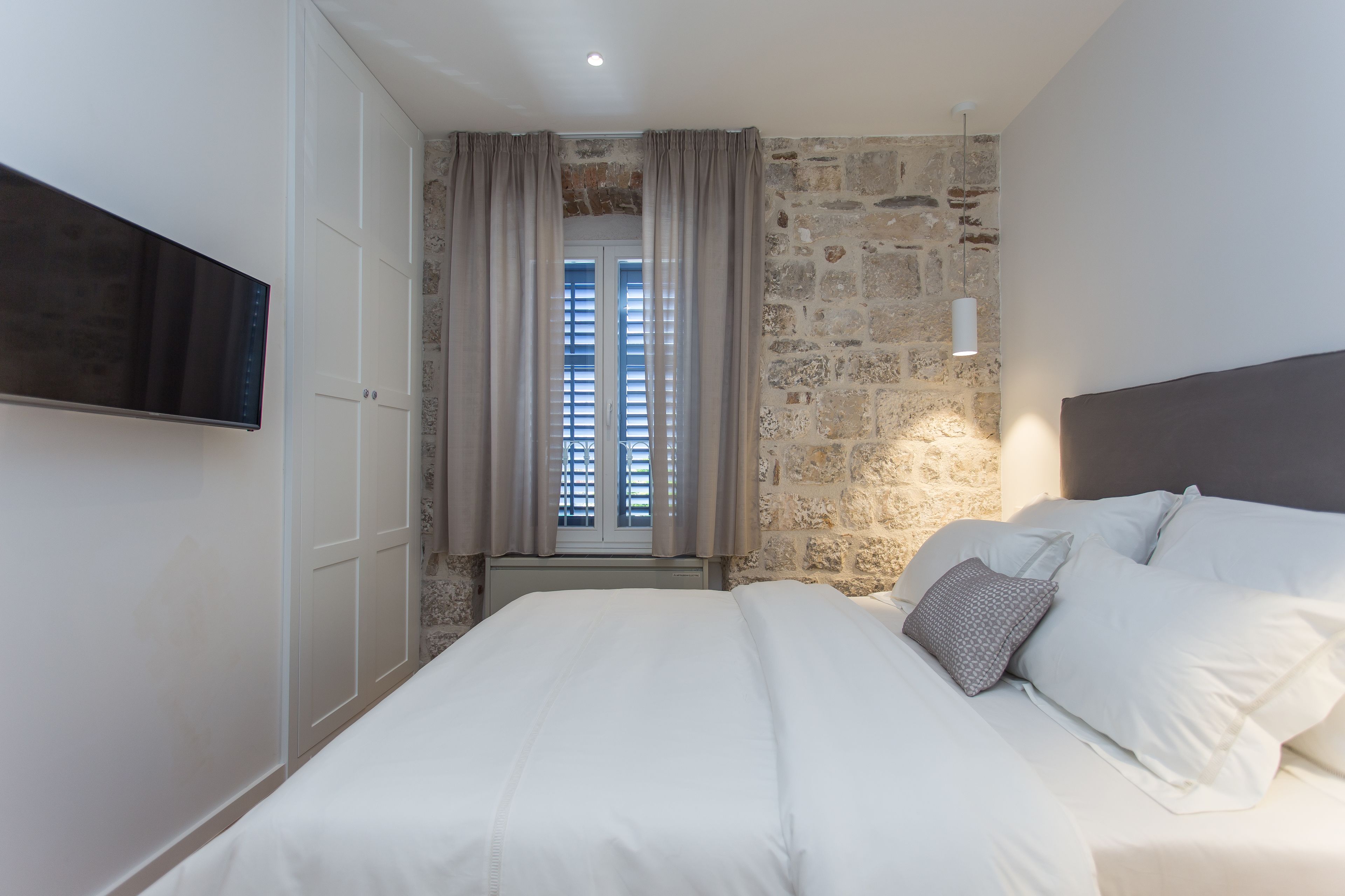 A bedroom with a stone wall, a bed with white bedding, a TV mounted on the wall, and a window with curtains.