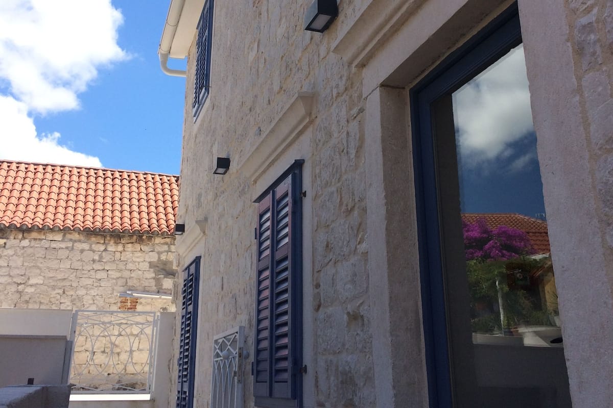 A stone building with a tiled roof, shutters, and a window with a view of purple flowers.