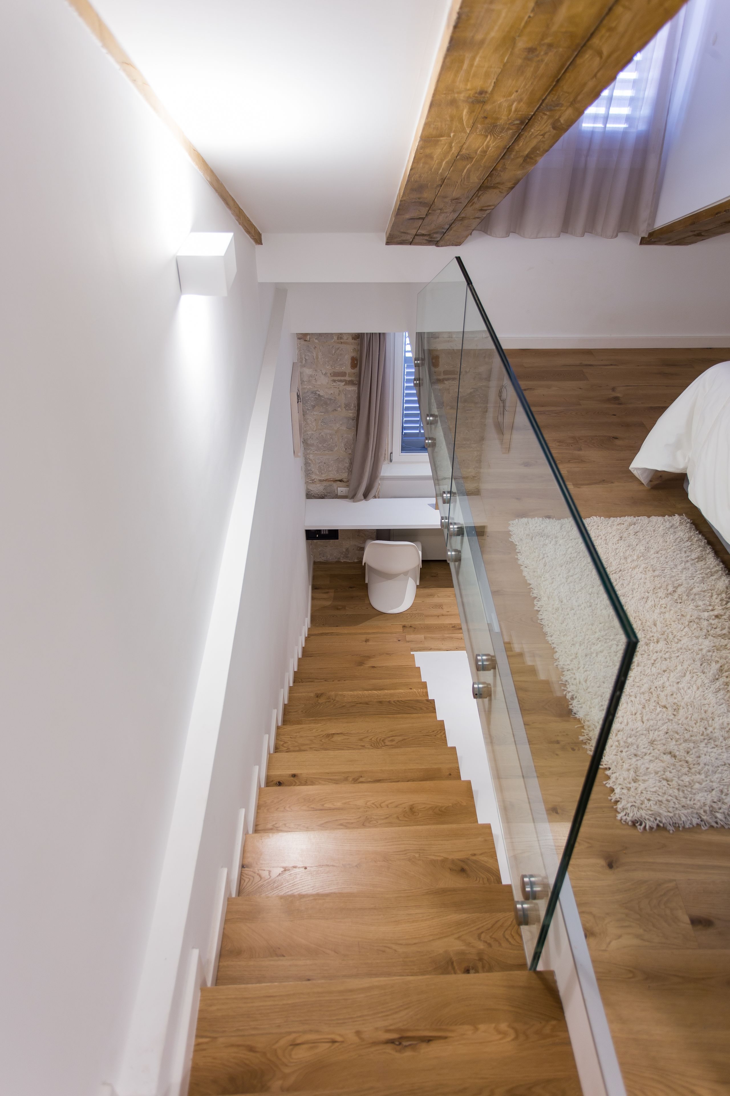Una escalera moderna con barandillas de vidrio que conduce a un dormitorio con cama blanca y alfombra blanca.