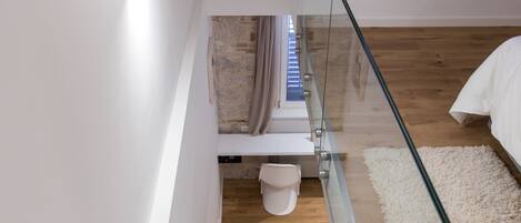 A modern staircase with glass railings leading to a bedroom with a white bed and a white rug.