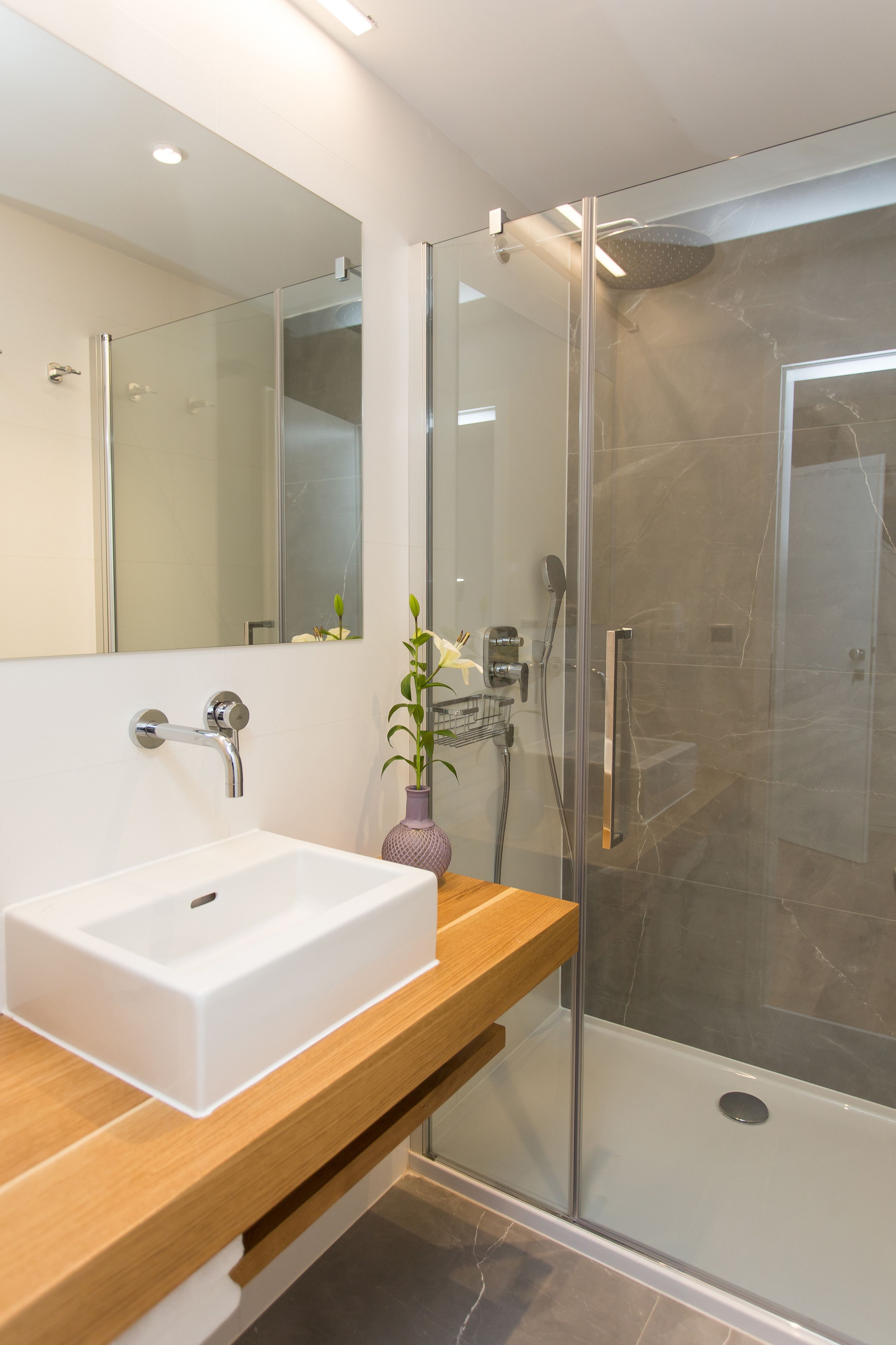 A modern bathroom with a white sink, a wooden vanity, a glass shower enclosure, and a large mirror.