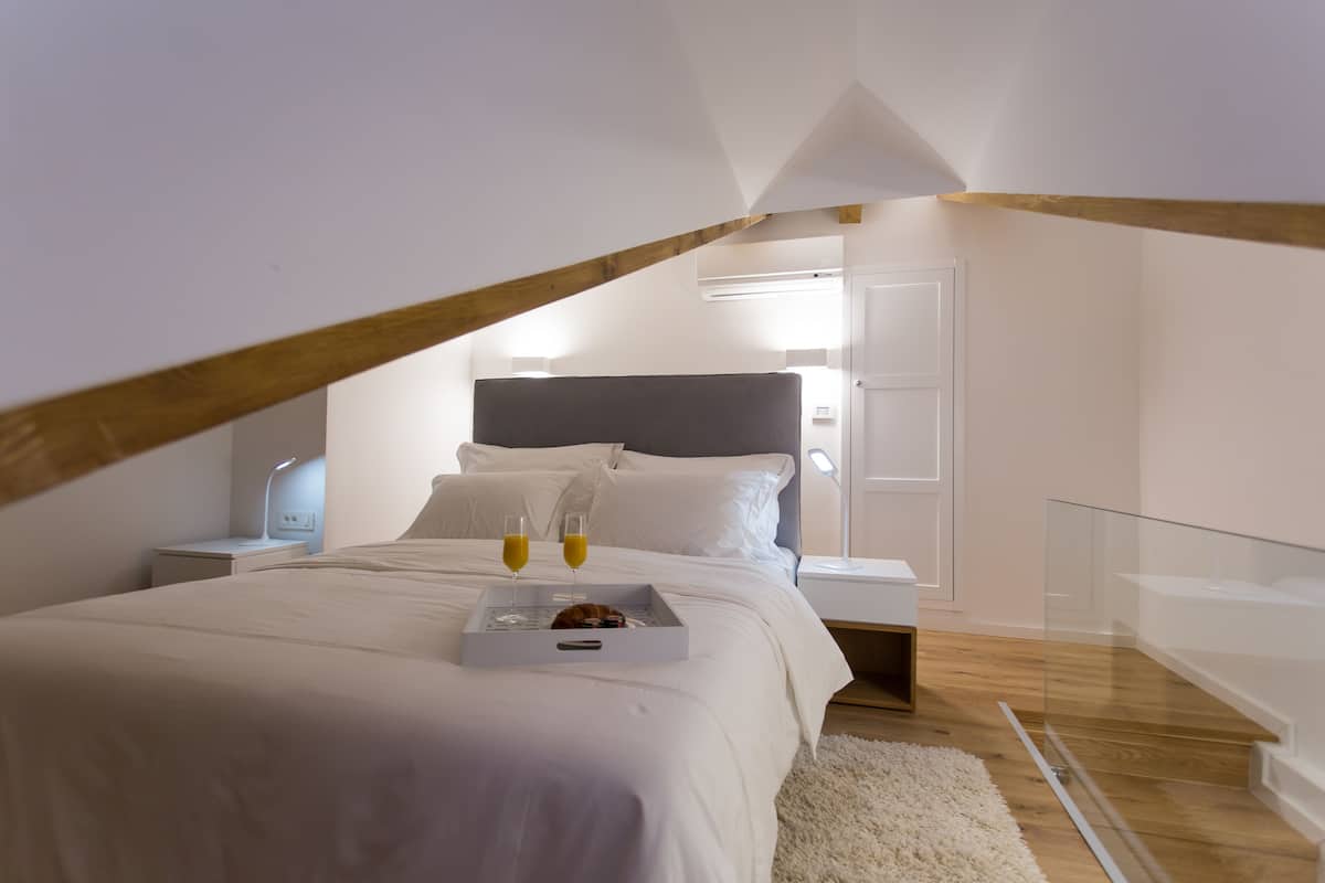 A modern bedroom with a sloped ceiling, a large bed with white bedding, a bedside table with a lamp, and a tray with two glasses of orange juice.