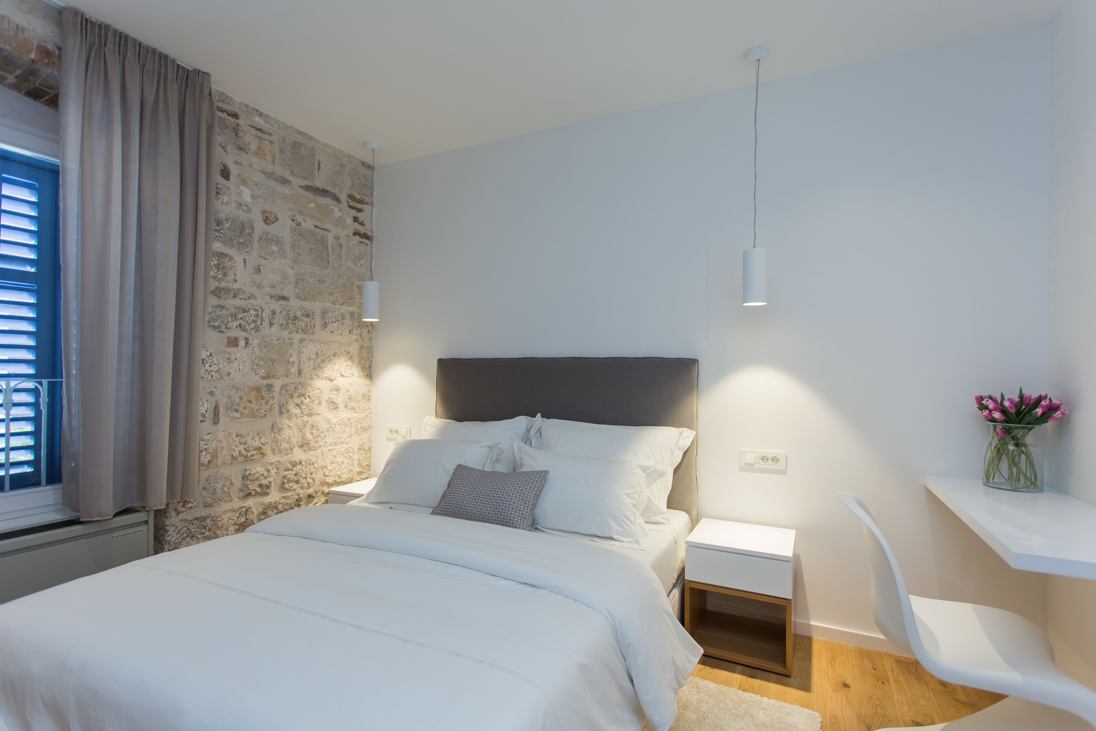 A modern bedroom with a stone wall, a large bed, a bedside table, a desk, and a hanging light fixture.